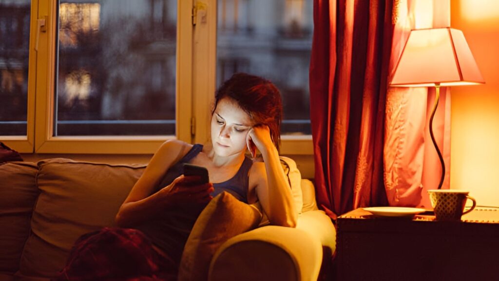 A woman sitting on a couch in a dimly lit living room at night, illuminated by the glow of her smartphone and a warm lamp. Her contemplative and tired expression reflects the post-holiday slump and the emotional transition back into the workweek.